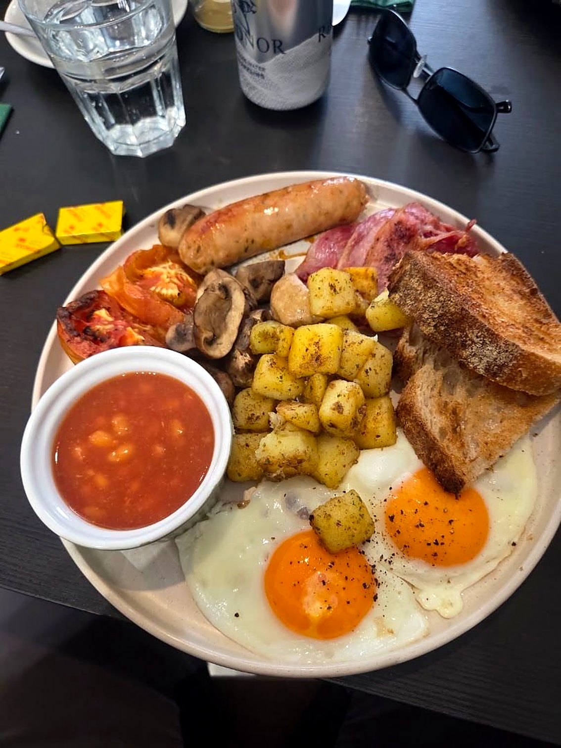 A full breakfast on a round plate: two fried eggs, sausage, bacon, roast potatoes, tomato, mushrooms, toast and a pot of beans