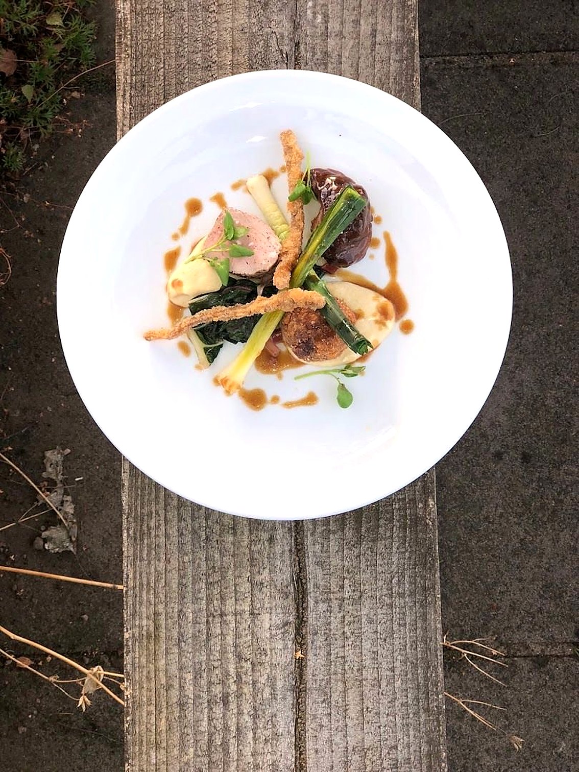 A plated dish on a white plate: sliced meat with greens, a light sauce and crisp shallots, set on a wooden plank